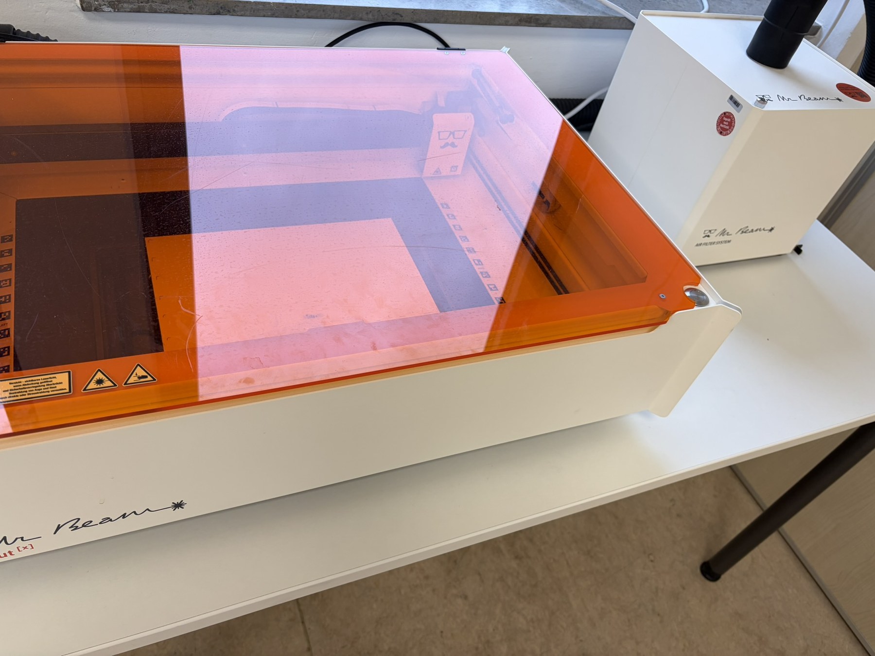 Lasercutter mit orangefarbener Schutzabdeckung und Absaug- und Filtereinheit